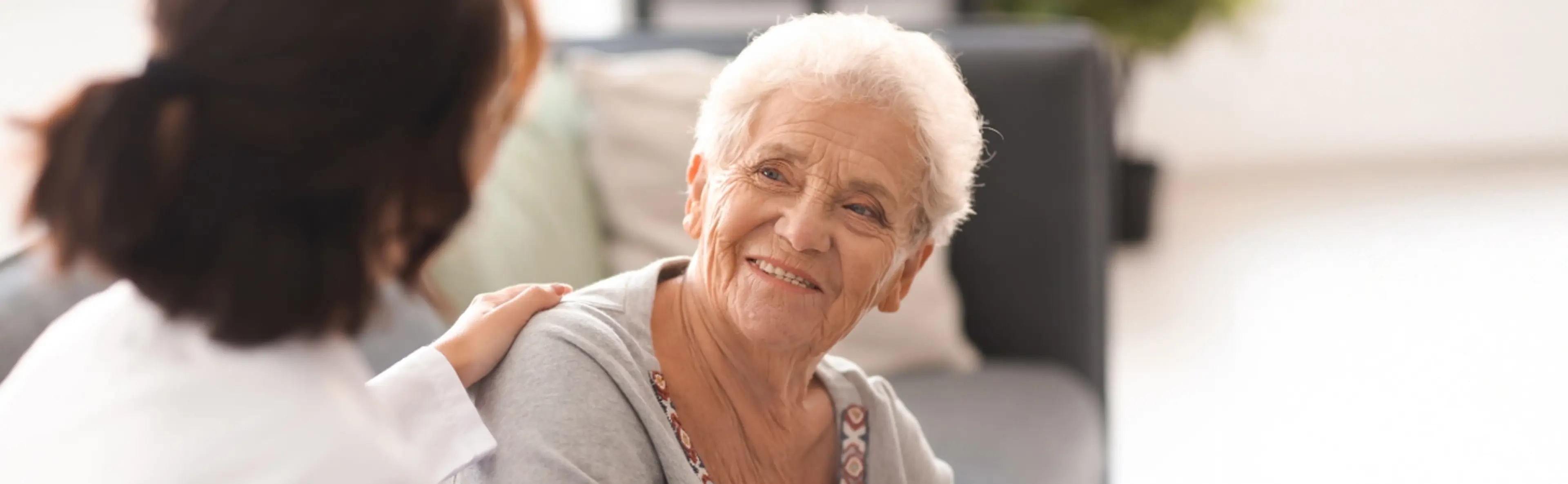 Une professionnelle de santé accompagne avec bienveillance une femme âgée souriante assise sur un canapé.
