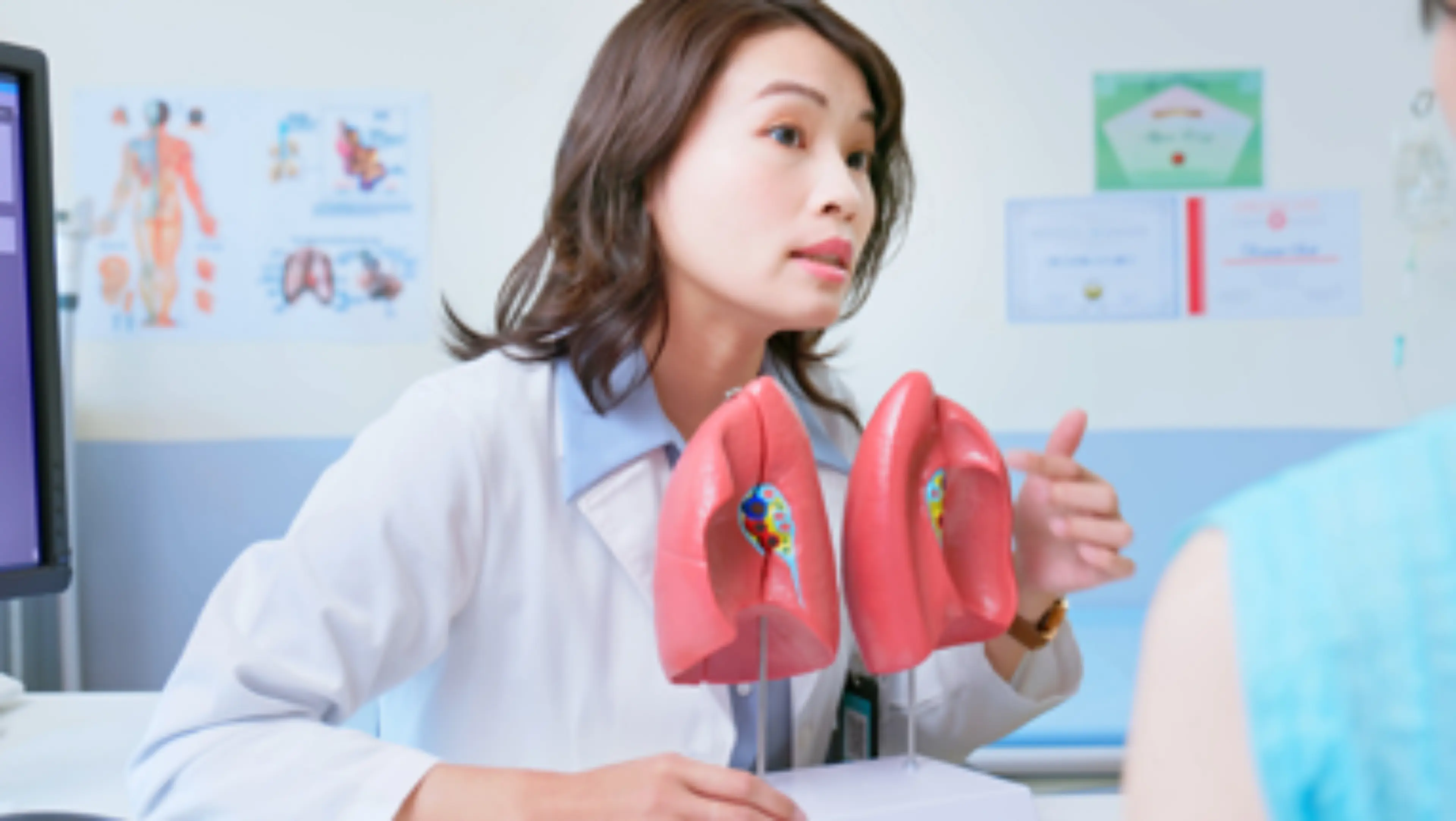 Consultation médicale : une professionnelle de santé en blouse blanche explique des résultats d'imagerie médicale à une patiente, avec des modèles anatomiques liées au cancer du sein sur le bureau.