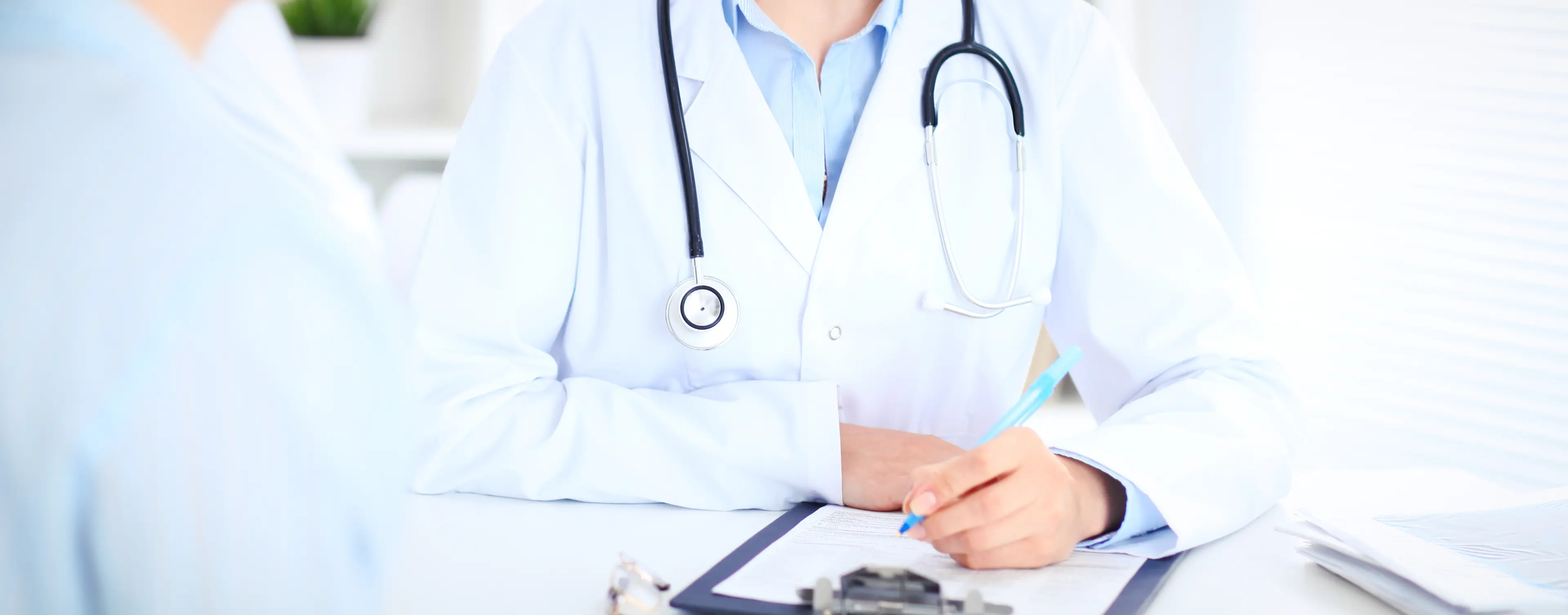 Arzt in weißem Kittel mit Stethoskop um den Hals sitzt an Schreibtisch und hält Stift über Klemmbrett mit Dokumenten