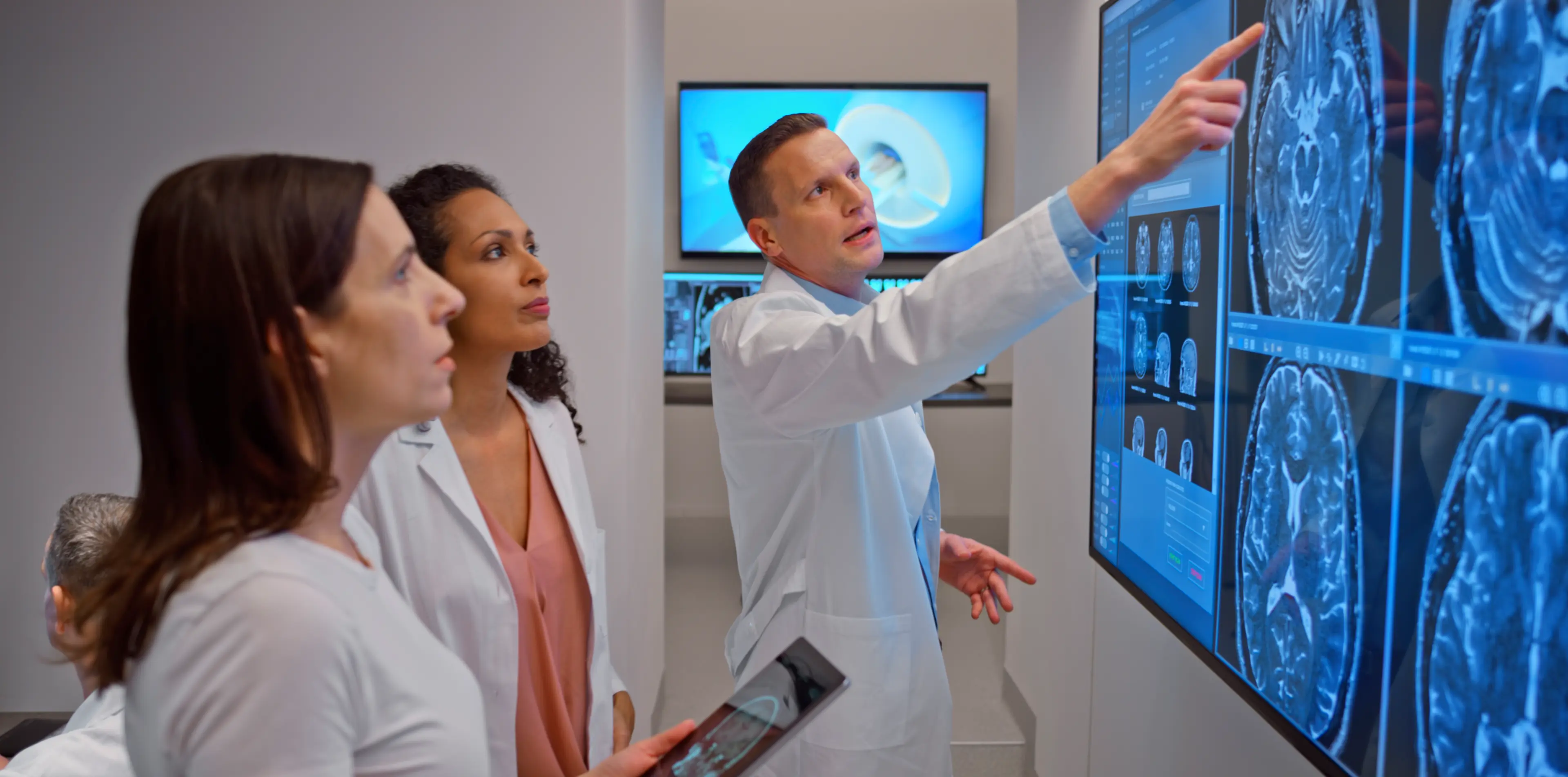 Drei Personen in Arztkleidung betrachten und besprechen medizinische Bildgebungen auf einem großen Bildschirm in einem Untersuchungsraum