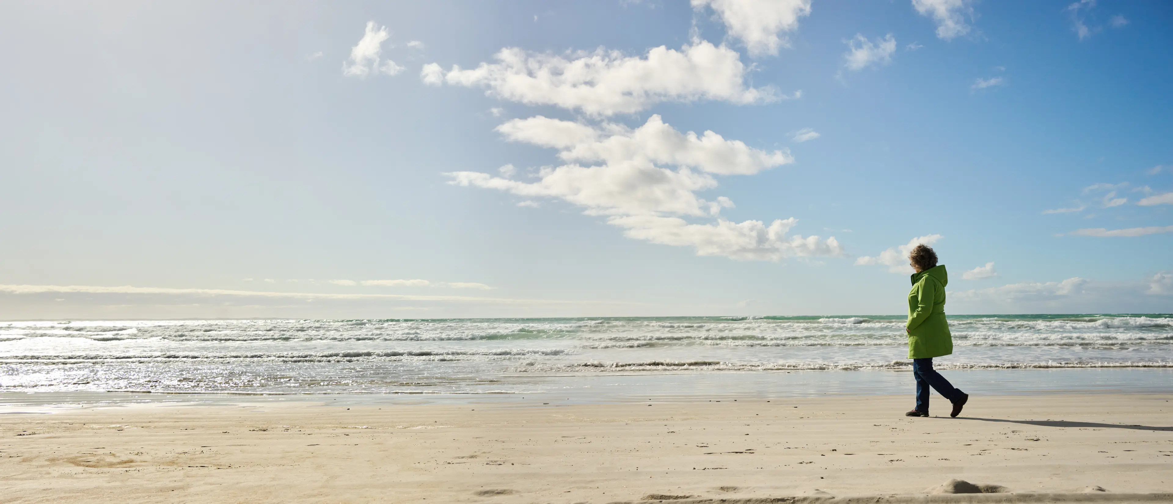 Person mit grüner Jacke geht am Strand entlang bei bewölktem Himmel