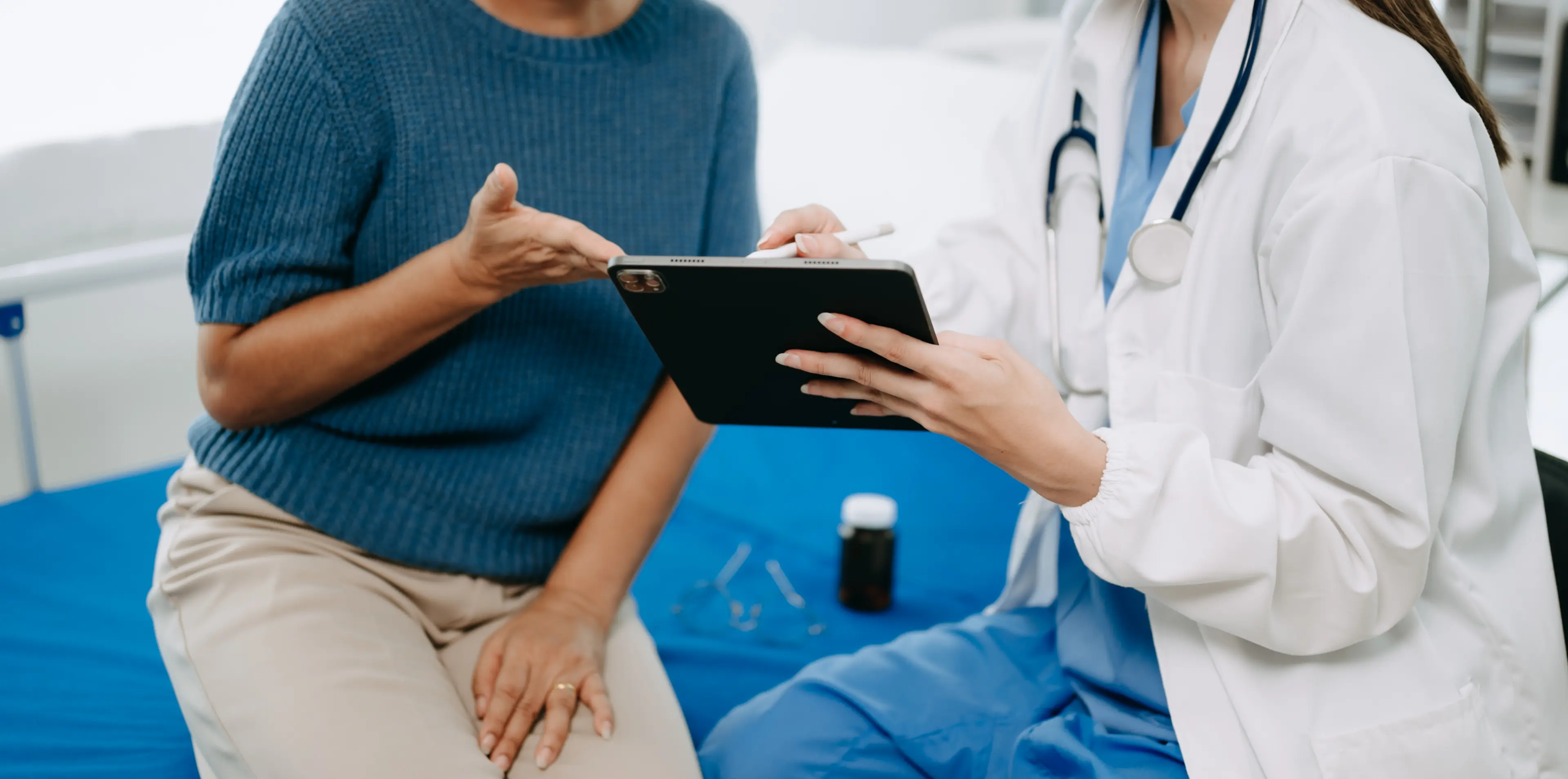 Ärztin mit Kittel und Stethoskop hält Tablet und spricht mit sitzender Patientin in blauem Pullover und beiger Hose