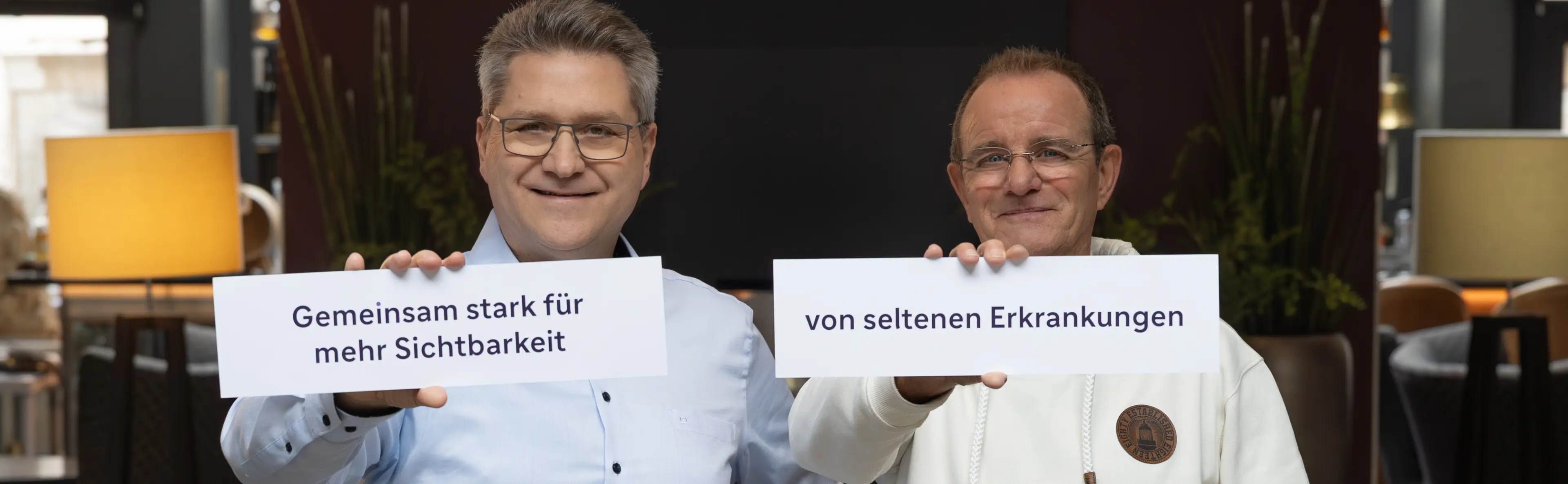 Zwei männliche Personen halten jeder lachend ein Schild vor sich. Jedes Schild enthält Bestandteile eines gemeinsamen Satzes. Zusammen ergibt es den Satz: „Gemeinsam stark für mehr Sichtbarkeit von seltenen Erkrankungen“