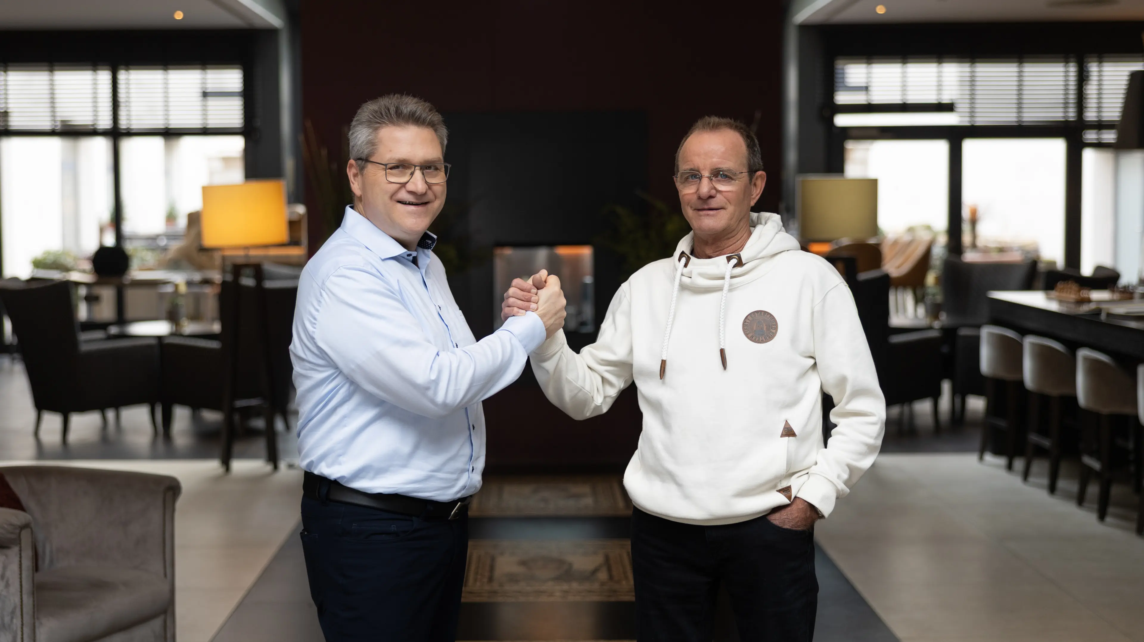 Zwei Männer stehen in einem modernen Innenraum und geben sich die Hand in einem festen, ineinander verschränkten Handschlag.