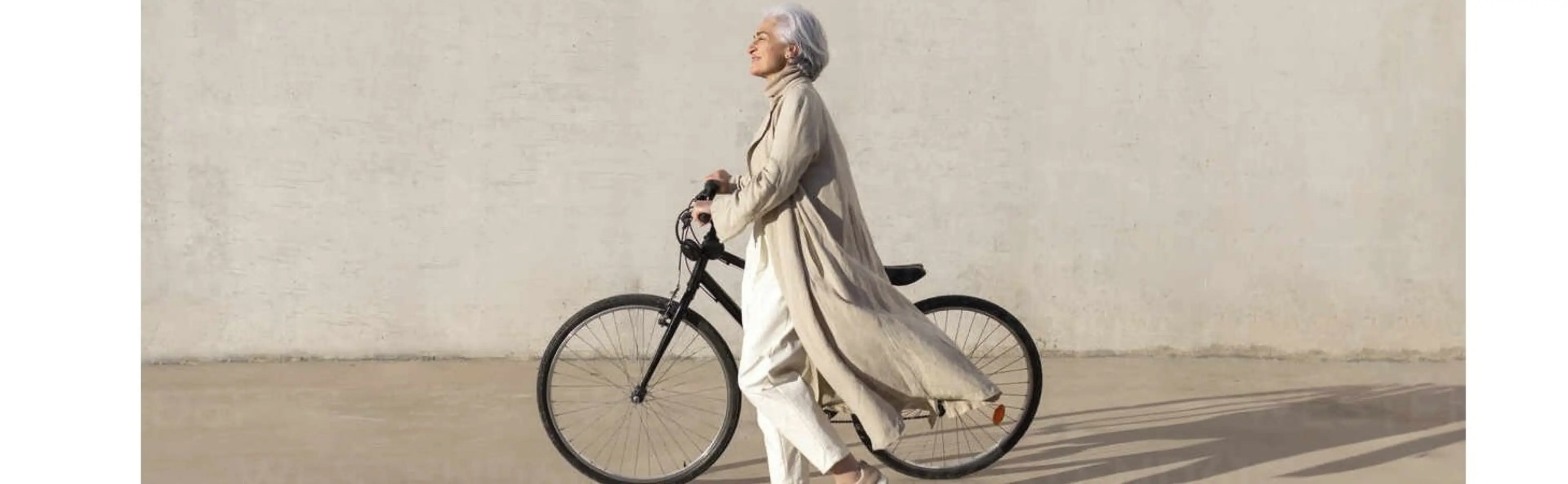 Eine reife Frau geht bei hellem Sonnenschein entspannt auf einem Gehweg und schiebt ihr Fahrrad an ihrer Seite.