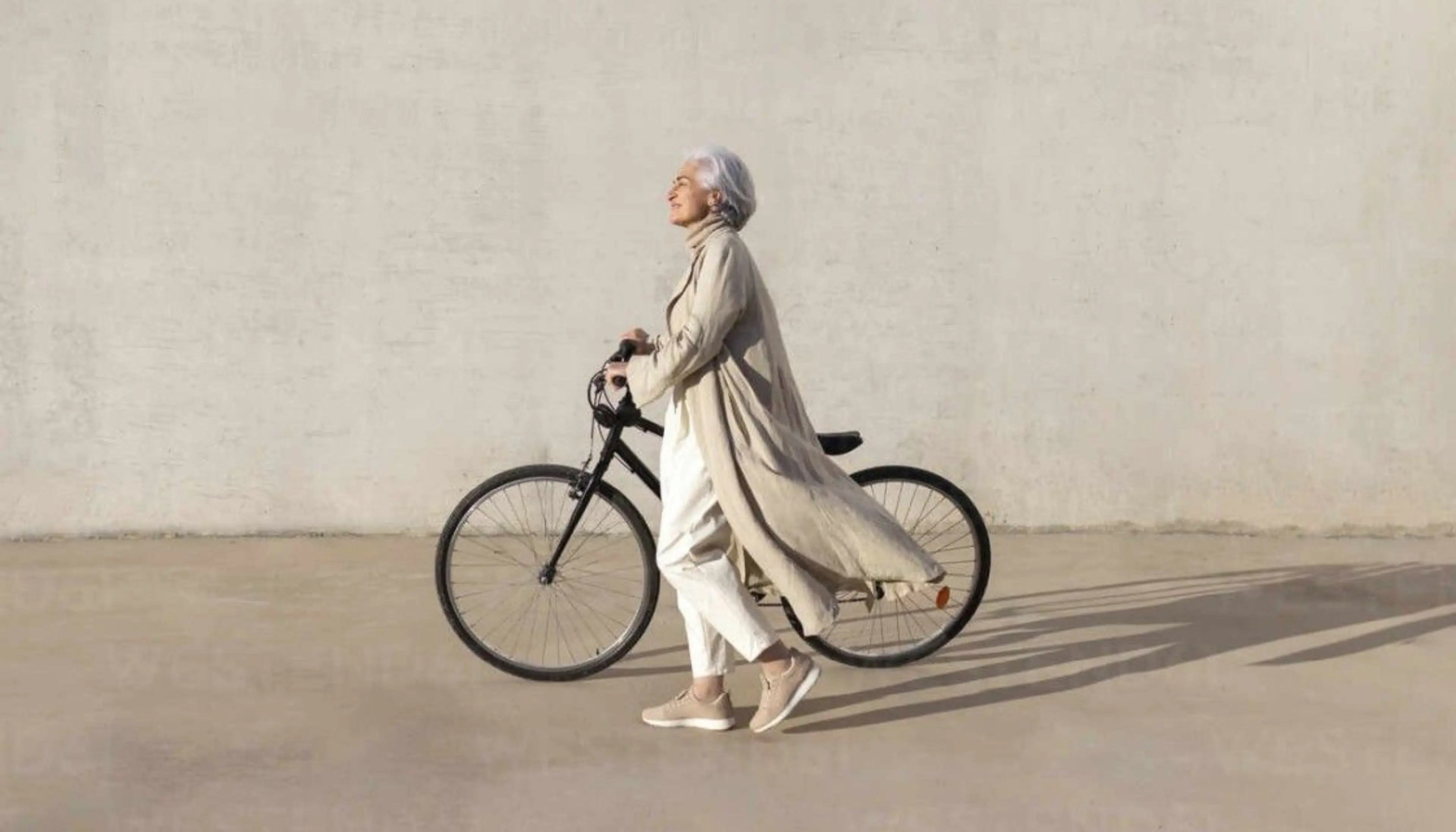 Eine reife Frau geht bei hellem Sonnenschein entspannt auf einem Gehweg und schiebt ihr Fahrrad an ihrer Seite.