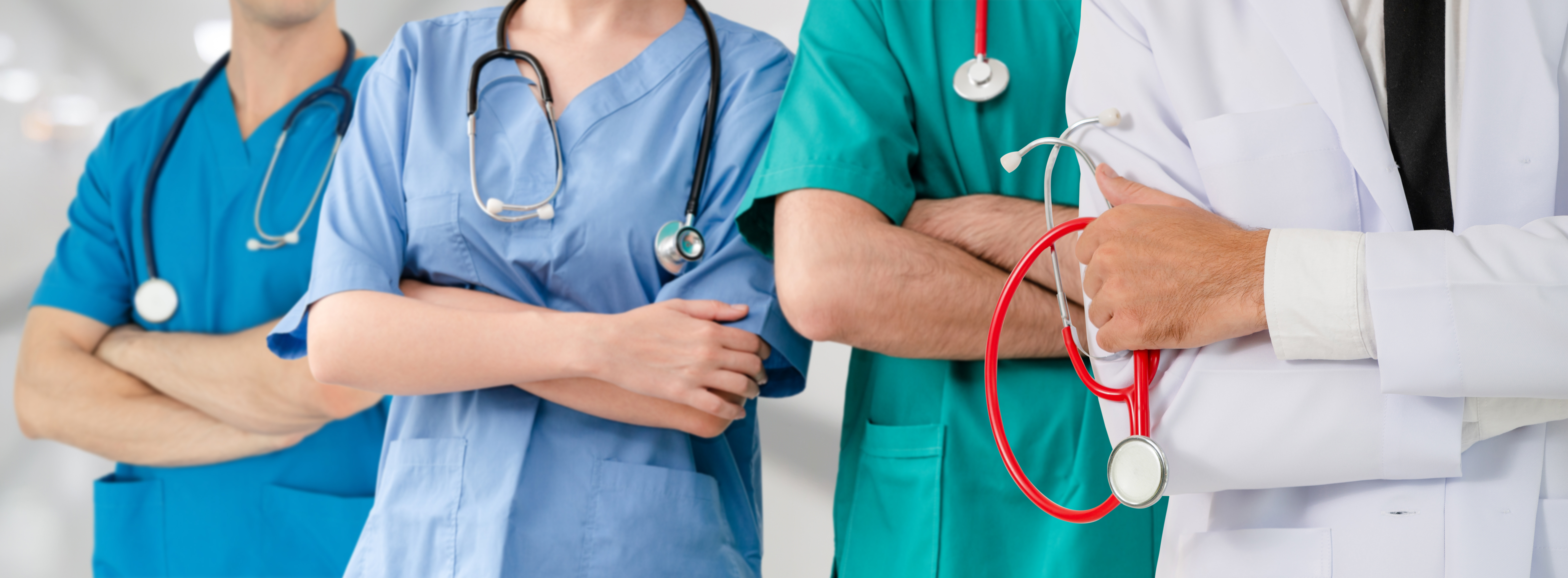 Gruppe von vier medizinischen Fachkräften mit verschränkten Armen, zwei tragen blaue, eine grüne und eine weiße Arbeitskleidung, zwei mit Stethoskopen um den Hals