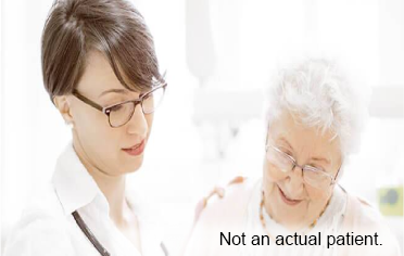 A healthcare professional wearing glasses places her hand on the shoulder of a elderly patient as they engage in conversation.