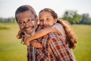 Elderly man wearing glasses gives a piggyback ride to a girl with long curly hair in an outdoor setting with green fields and trees in the background.