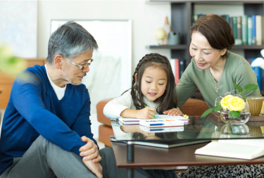 Two adults and a child are sitting together at a table in a living room, engaged in a writing activity, with bookshelves, plants, and decorative items in the background.