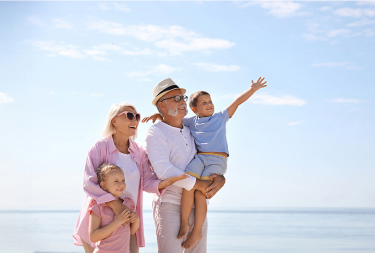Family of four enjoying time together, a woman in a pink jacket with a girl in a pink shirt, and a man wearing a hat and glasses carrying a boy in a blue shirt, with the ocean and sky in the background.