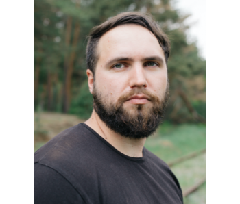 Junger Mann mit dunklem T-Shirt und kurzem Bart steht vor unscharfem Waldhintergrund.