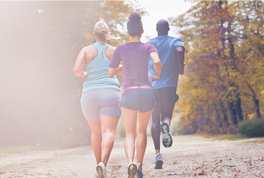 Two women and one man running on a forest trail with autumn leaves and sunlight filtering through the trees