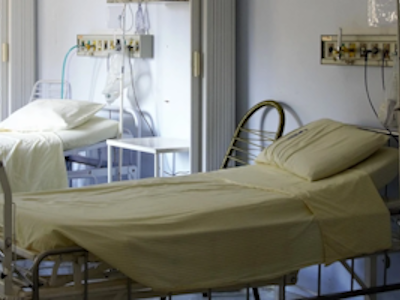 Zwei Krankenhausbetten mit gelben Bettlaken und Kissen in einem Patientenzimmer, medizinische Geräte an der Wand.