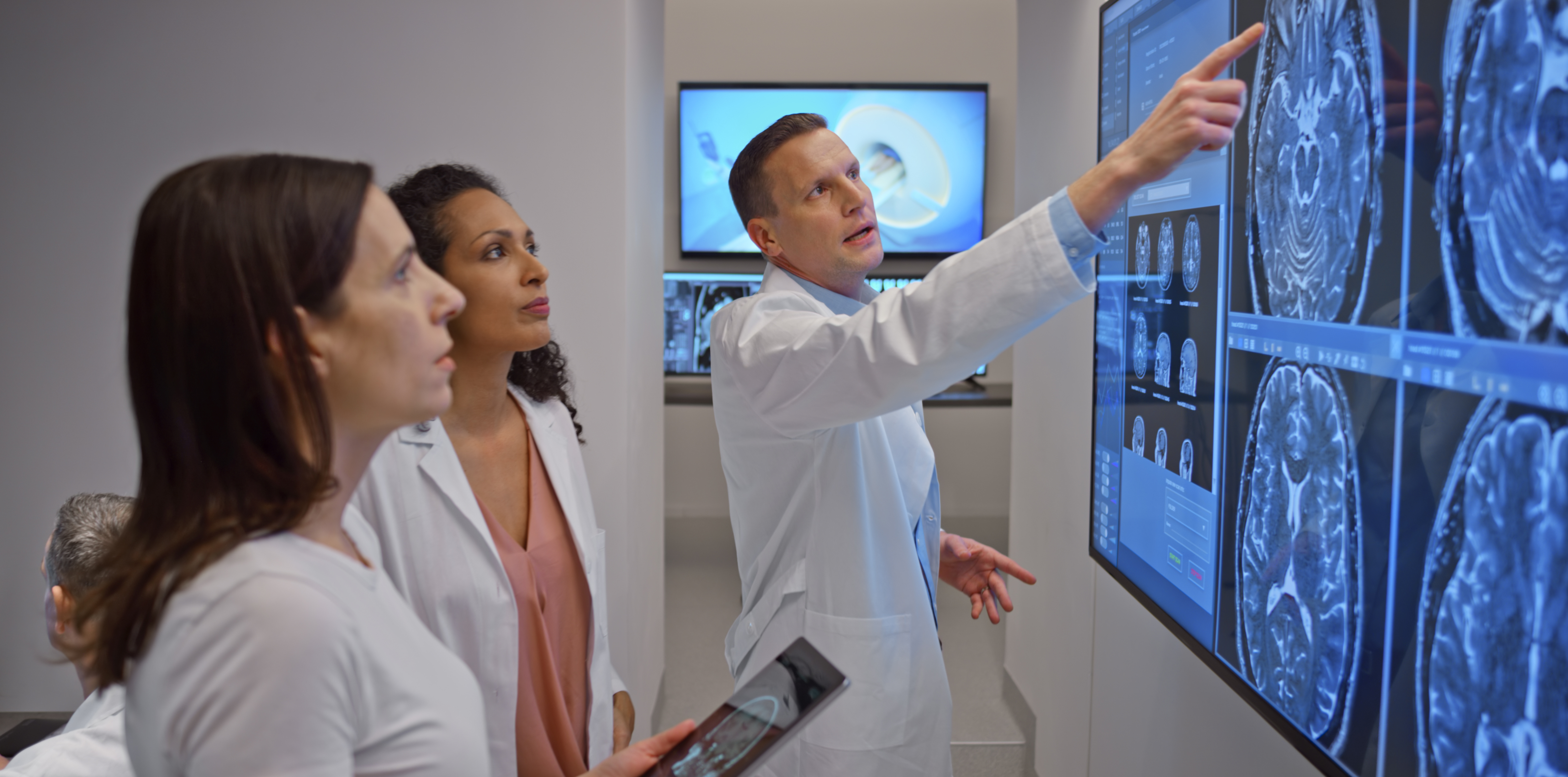 Drei Personen in Arztkleidung betrachten und besprechen medizinische Bildgebungen auf einem großen Bildschirm in einem Untersuchungsraum