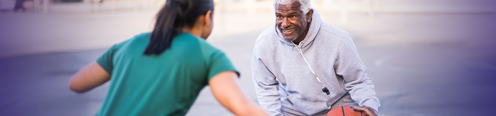 A smiling elderly man wearing a gray hoodie, with a black whistle around his neck, is playing with a girl in a green T-shirt.
