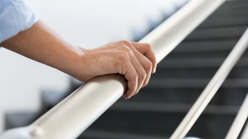 Hand greift einen weißen Handlauf an einer Treppe, unscharfer Hintergrund mit Treppenstufen.